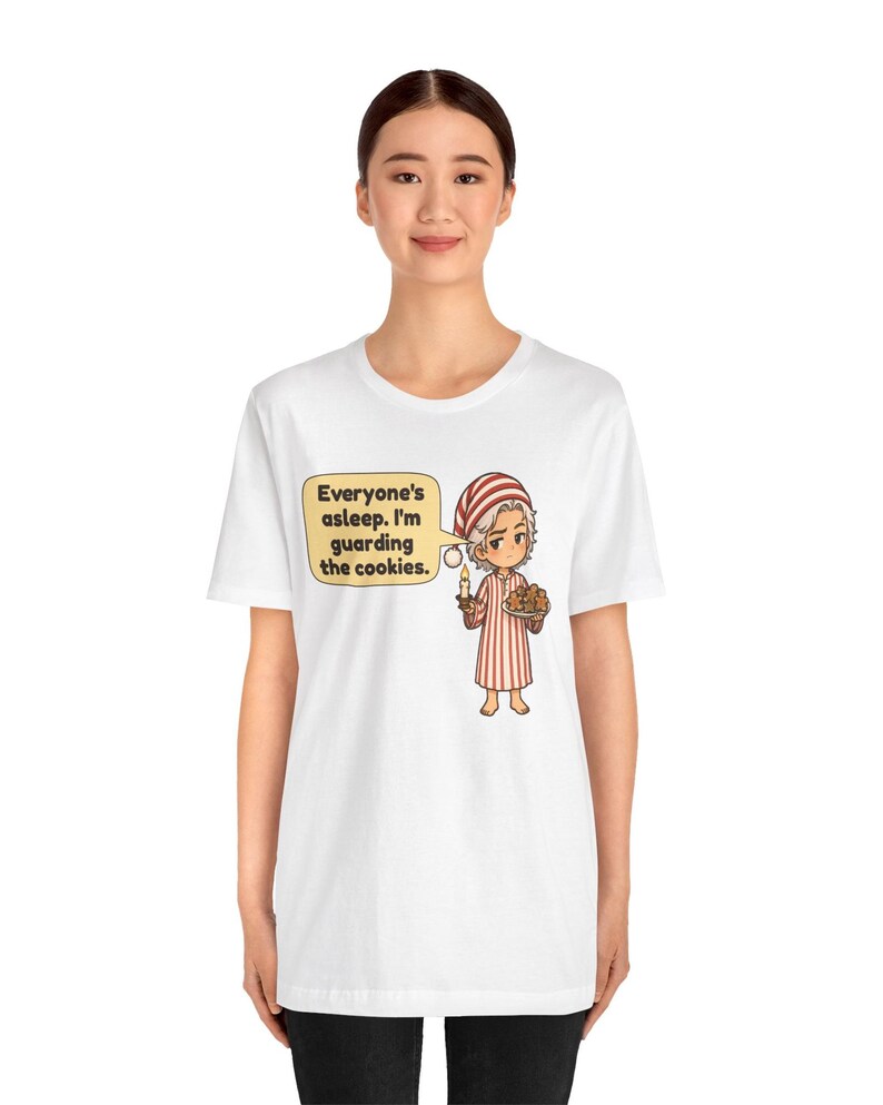 Op de afbeelding: Een wit T-shirt met korte mouwen en een cartoonillustratie van een figuur in een rood-wit gestreepte pyjama die een kaars en een bord koekjes vasthoudt. Een tekstballon bevat de tekst: Everyone's asleep. I'm guarding the cookies.