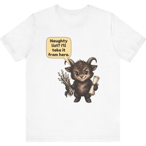 Puede incluir: Una camiseta blanca con el gr&aacute;fico de una criatura marr&oacute;n con cuernos que sostiene un manojo de palos y un pergamino. Un globo de texto sobre la criatura contiene el texto: Naughty list? I'll take it from here. El dise&ntilde;o est&aacute; centrado.