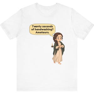 Op de afbeelding: Een wit t-shirt met een cartoonillustratie van een persoon met lang bruin haar in een cr&egrave;mekleurige jurk en donkere sjaal, die een kaars vasthoudt. Een tekstballon erboven bevat de tekst: Twenty seconds of handwashing? Amateurs.