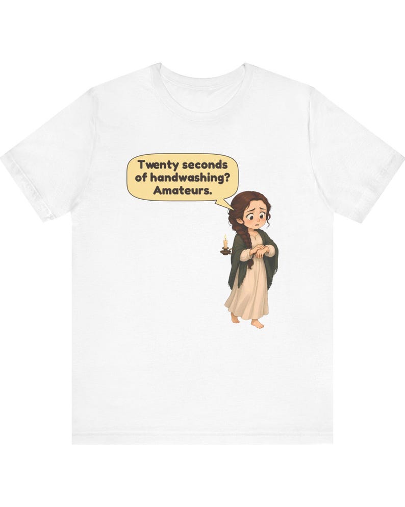 Op de afbeelding: Een wit t-shirt met een cartoonillustratie van een persoon met lang bruin haar in een cr&egrave;mekleurige jurk en donkere sjaal, die een kaars vasthoudt. Een tekstballon erboven bevat de tekst: Twenty seconds of handwashing? Amateurs.