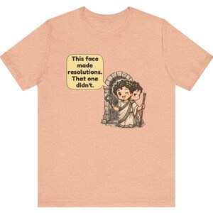 K&ouml;nnte beinhalten: Ein pfirsichfarbenes T-Shirt mit einer Grafik des zweigesichtigen r&ouml;mischen Gottes Janus, der einen Lorbeerkranz tr&auml;gt. Ein Gesicht zwinkert und h&auml;lt einen Schl&uuml;ssel, das andere wirkt streng. Eine Sprechblase enth&auml;lt den Text: This face made resolutions. That one didn't.