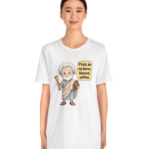 May include: A white t-shirt featuring a cartoon illustration of a bearded man in a toga holding a scroll and a brown jug. A speech bubble next to him contains the text: First, do no harm. Second, coffee. The design is centered on the chest.