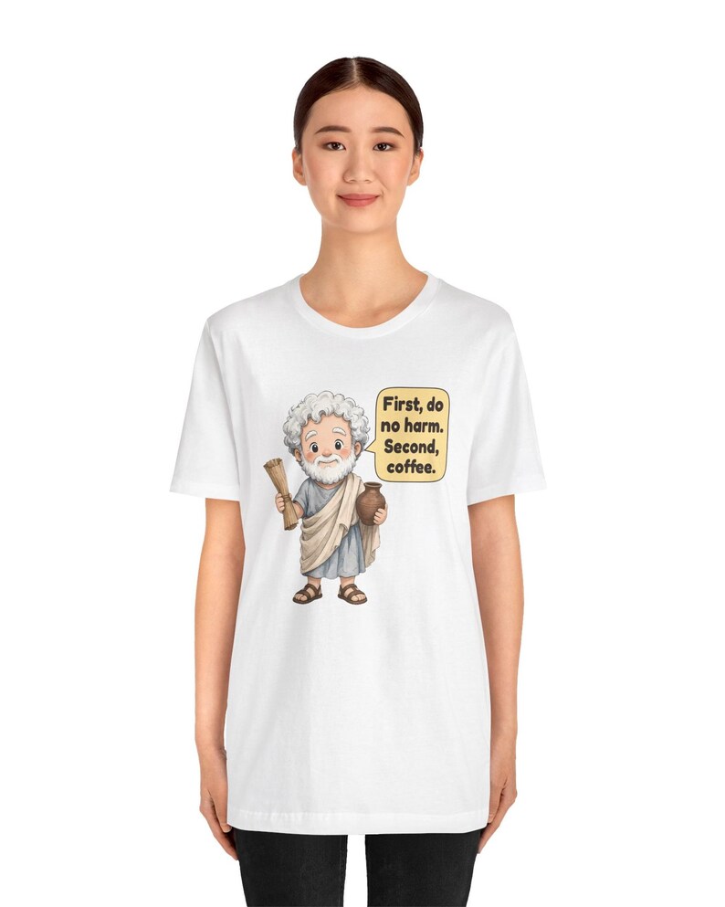May include: A white t-shirt featuring a cartoon illustration of a bearded man in a toga holding a scroll and a brown jug. A speech bubble next to him contains the text: First, do no harm. Second, coffee. The design is centered on the chest.