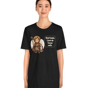 May include: A black t-shirt featuring a graphic of a cartoon angel with curly brown hair, white wings, and a red cloak, holding a wooden staff and a fish. A speech bubble next to the angel contains the text: God heals. I just do house calls.