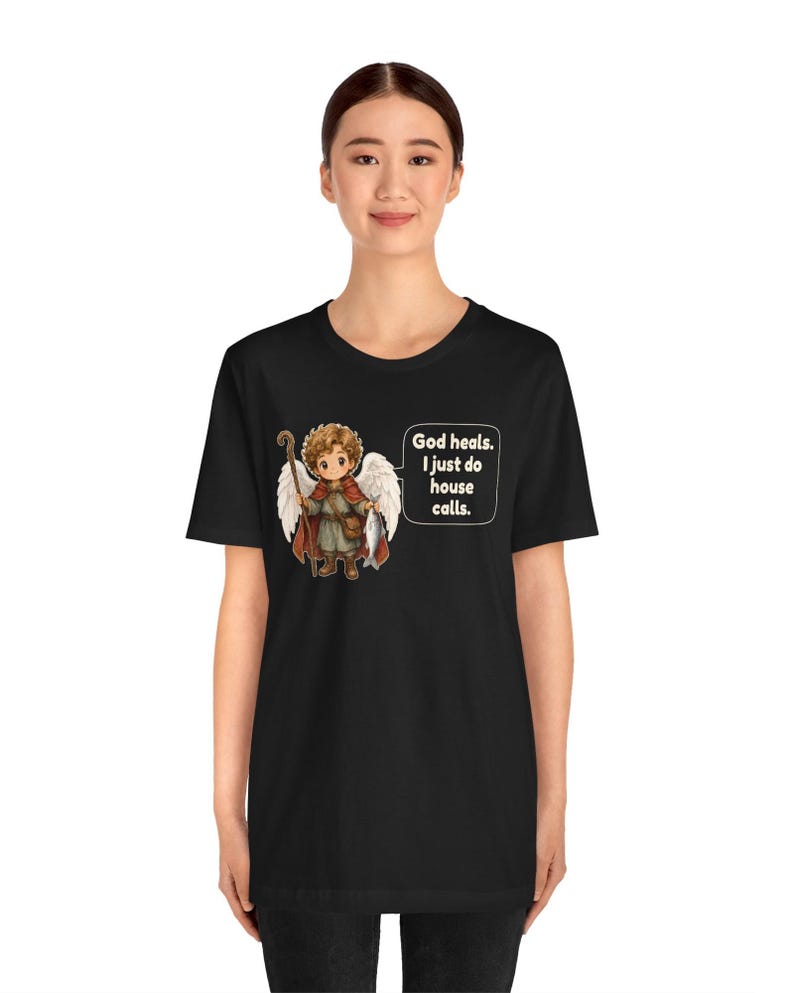 May include: A black t-shirt featuring a graphic of a cartoon angel with curly brown hair, white wings, and a red cloak, holding a wooden staff and a fish. A speech bubble next to the angel contains the text: God heals. I just do house calls.