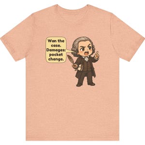 May include: A peach-colored t-shirt featuring a cartoon illustration of a historical figure in a brown suit holding a quill. A speech bubble contains the text: Won the case. Damages: pocket change. The design is centered on the chest.