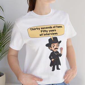 Pu&ograve; includere: Una t-shirt bianca a maniche corte con un'illustrazione a cartoni animati di un uomo in abito e cappello neri che tiene in mano un mazzo di carte. Un fumetto giallo sopra il personaggio contiene il testo: Thirty seconds of fame. Fifty years of interviews.