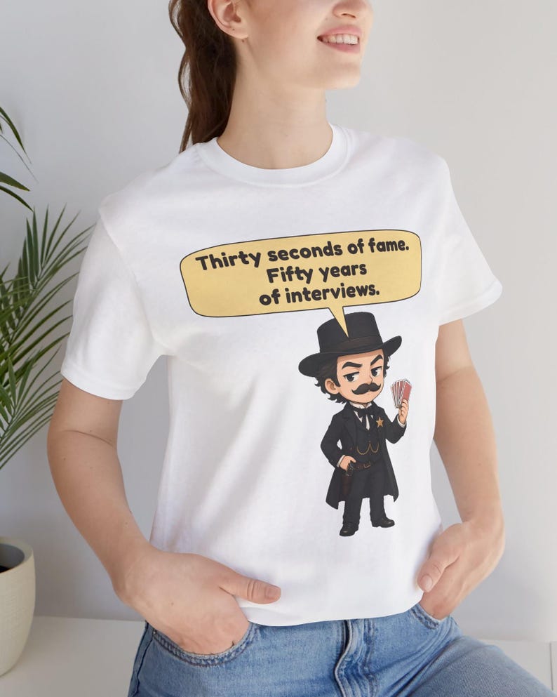 Pu&ograve; includere: Una t-shirt bianca a maniche corte con un'illustrazione a cartoni animati di un uomo in abito e cappello neri che tiene in mano un mazzo di carte. Un fumetto giallo sopra il personaggio contiene il testo: Thirty seconds of fame. Fifty years of interviews.