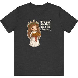 May include: A dark gray t-shirt featuring a cartoon illustration of a person wearing a white robe and a candle crown, holding a tray of buns. A speech bubble next to the figure contains the text: Bringing the light (and the buns).