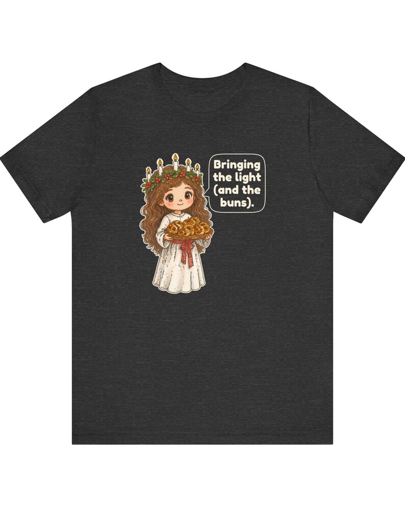 May include: A dark gray t-shirt featuring a cartoon illustration of a person wearing a white robe and a candle crown, holding a tray of buns. A speech bubble next to the figure contains the text: Bringing the light (and the buns).