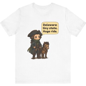 May include: A white t-shirt featuring a cartoon illustration of a person in a colonial-style hat and coat standing next to a small brown horse. A speech bubble above them contains the text: Delaware: tiny state. Huge ride.