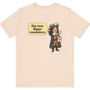 May include: A light beige t-shirt featuring a cartoon illustration of a historical figure with a large nose, wearing a hat with a feather, a cape, and holding a quill and letter. A speech bubble contains the text: Big nose. Bigger vocabulary.