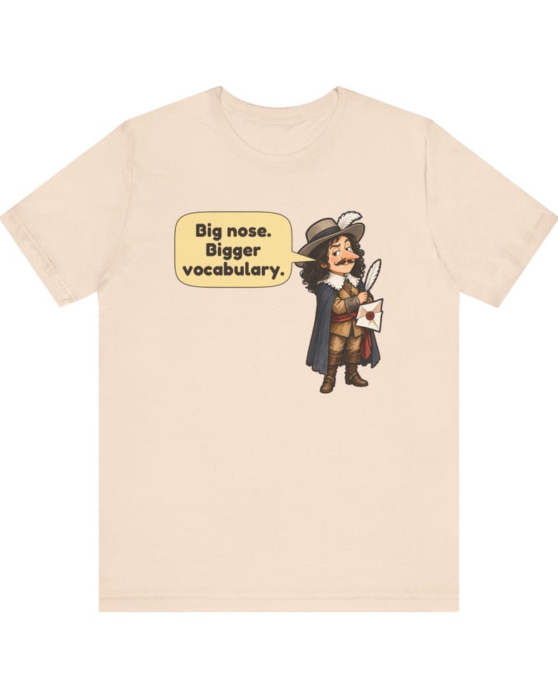 May include: A light beige t-shirt featuring a cartoon illustration of a historical figure with a large nose, wearing a hat with a feather, a cape, and holding a quill and letter. A speech bubble contains the text: Big nose. Bigger vocabulary.