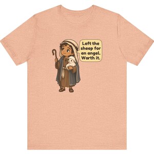 May include: A peach-colored t-shirt featuring a cartoon illustration of a shepherd holding a small white lamb and a wooden staff. A speech bubble next to the character contains the text: Left the sheep for an angel. Worth it.