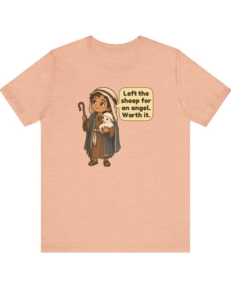 May include: A peach-colored t-shirt featuring a cartoon illustration of a shepherd holding a small white lamb and a wooden staff. A speech bubble next to the character contains the text: Left the sheep for an angel. Worth it.