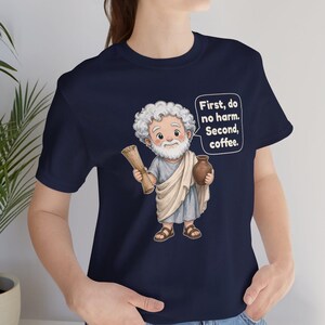 May include: A navy blue t-shirt featuring a cartoon illustration of a bearded figure in a light blue tunic and white toga, holding a scroll and a brown jug. A speech bubble contains the text: First, do no harm. Second, coffee.