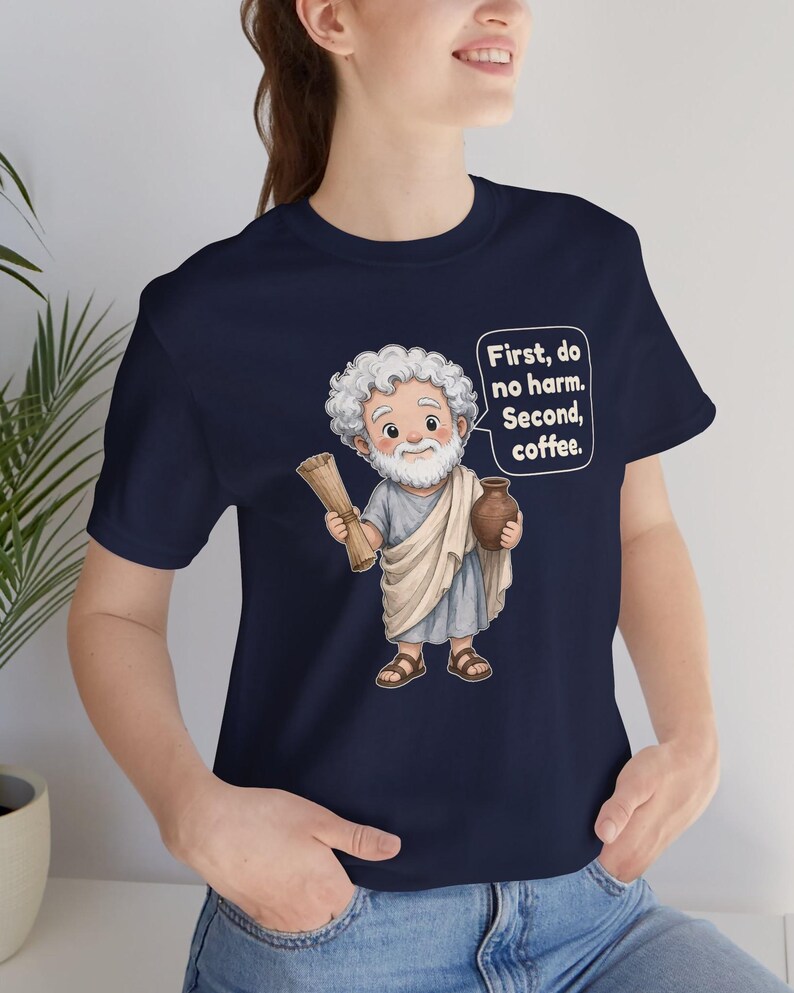 May include: A navy blue t-shirt featuring a cartoon illustration of a bearded figure in a light blue tunic and white toga, holding a scroll and a brown jug. A speech bubble contains the text: First, do no harm. Second, coffee.