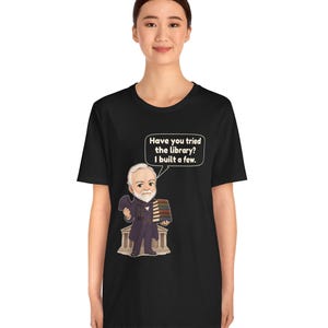 May include: A black t-shirt featuring a cartoon illustration of a bearded man in a suit holding a stack of books in front of a building. A speech bubble above him contains the text: Have you tried the library? I built a few.
