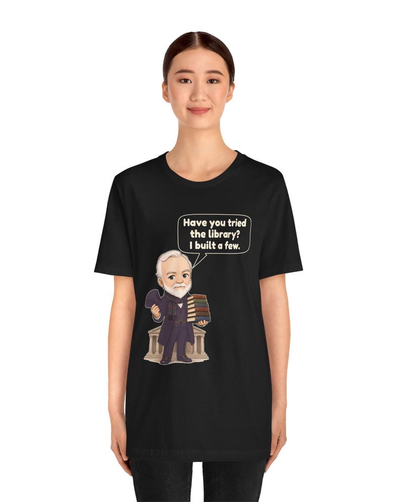 May include: A black t-shirt featuring a cartoon illustration of a bearded man in a suit holding a stack of books in front of a building. A speech bubble above him contains the text: Have you tried the library? I built a few.