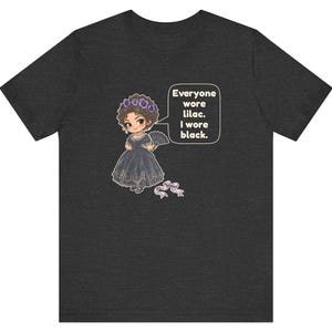 May include: A dark heather gray short-sleeve t-shirt featuring a cartoon character in a black dress with a lilac flower crown and fan. A speech bubble contains the text: Everyone wore lilac. I wore black. A small lilac ribbon sits below.