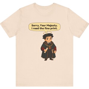 May include: A light beige t-shirt featuring a cartoon illustration of a person wearing a dark robe, a chain with a red pendant, and a halo. The person holds a scroll and a book. A speech bubble above contains the text: Sorry, Your Majesty. I read the fine print.