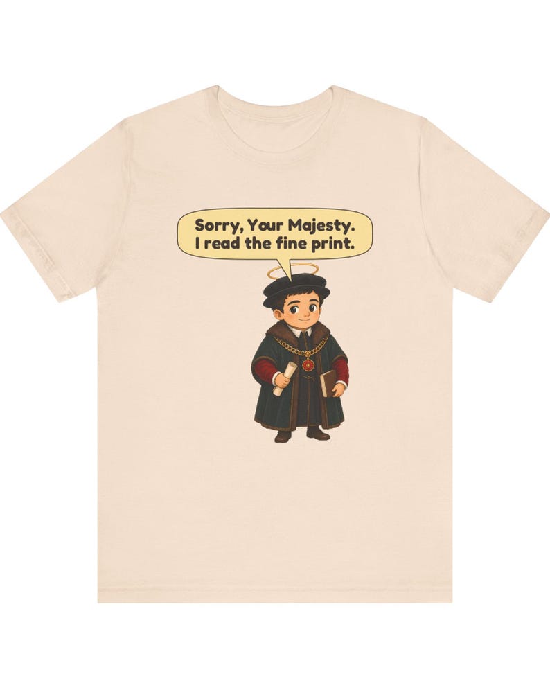 May include: A light beige t-shirt featuring a cartoon illustration of a person wearing a dark robe, a chain with a red pendant, and a halo. The person holds a scroll and a book. A speech bubble above contains the text: Sorry, Your Majesty. I read the fine print.