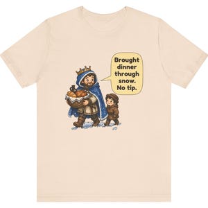May include: A light beige t-shirt featuring a cartoon illustration of a king in a blue cloak carrying a basket of food with a child. A speech bubble contains the text: Brought dinner through snow. No tip. The design is centered on the chest.