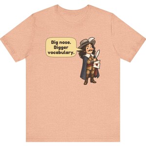 May include: A peach-colored t-shirt featuring a cartoon illustration of a historical figure with a large nose, wearing a hat and cape, holding a quill and letter. A speech bubble contains the text: Big nose. Bigger vocabulary.