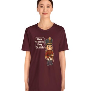 Pu&ograve; includere: Una t-shirt a maniche corte color bordeaux con la grafica di un soldatino schiaccianoci in uniforme rossa e un cappello alto. Accanto al soldato c'&egrave; un fumetto con il testo: Hard to crack. Easy to love.