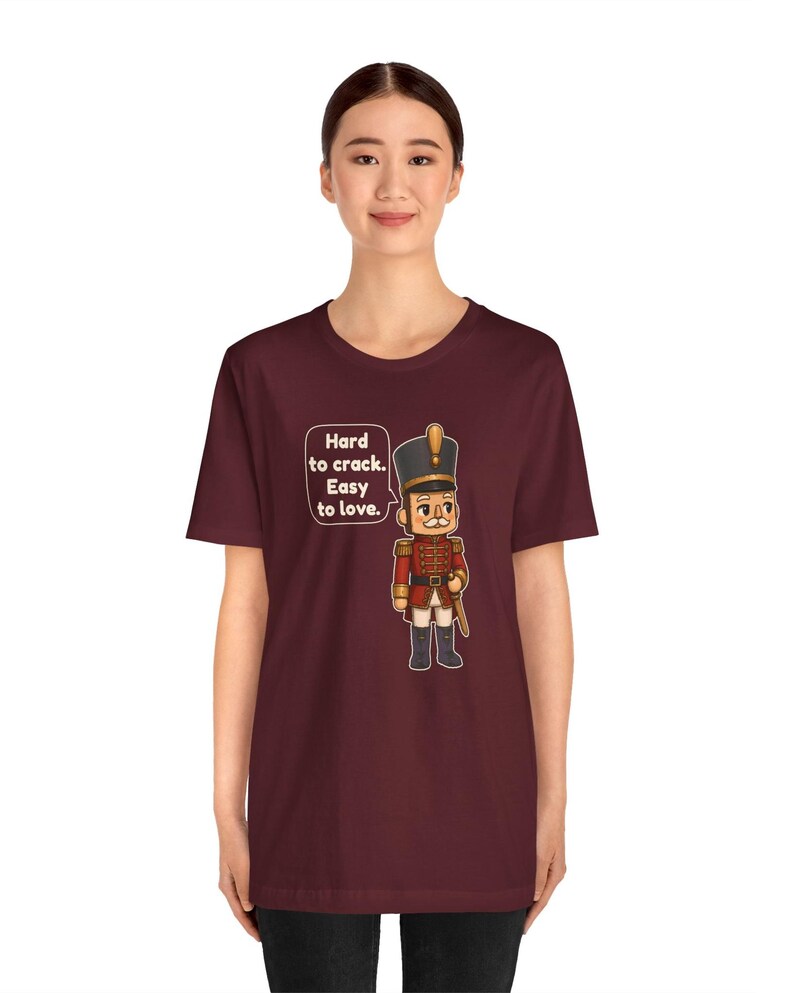 Pu&ograve; includere: Una t-shirt a maniche corte color bordeaux con la grafica di un soldatino schiaccianoci in uniforme rossa e un cappello alto. Accanto al soldato c'&egrave; un fumetto con il testo: Hard to crack. Easy to love.