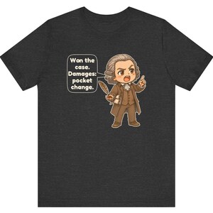 May include: A dark heather gray t-shirt featuring a cartoon illustration of a lawyer in a brown suit holding a quill. A speech bubble next to the character contains the text: Won the case. Damages: pocket change.