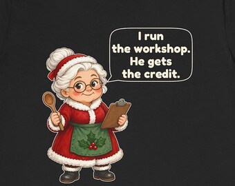 May include: A black t-shirt featuring a cartoon illustration of an elderly woman with white hair in a bun, wearing a red dress with white fur trim, a green apron with holly berries, and glasses. She holds a wooden spoon and clipboard.