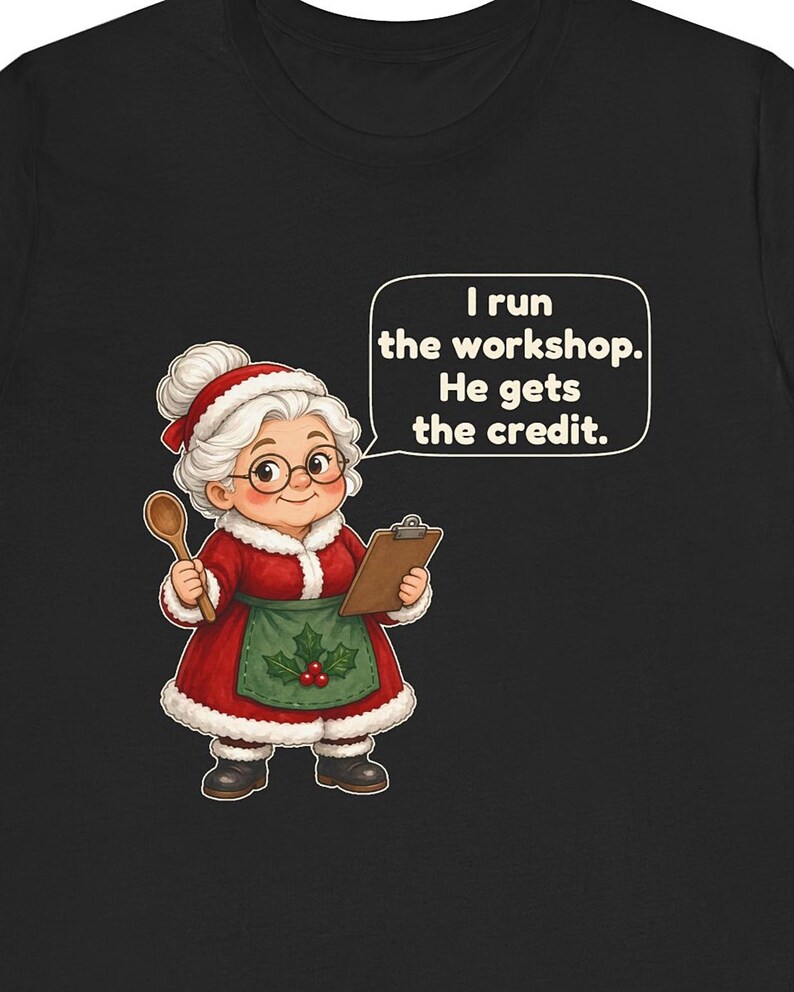 K&ouml;nnte beinhalten: Ein schwarzes T-Shirt mit einer Cartoon-Illustration einer &auml;lteren Frau mit wei&szlig;em Dutt, die ein rotes Kleid mit wei&szlig;em Pelzbesatz, eine gr&uuml;ne Sch&uuml;rze mit Stechpalmenbeeren und eine Brille tr&auml;gt. Sie h&auml;lt einen Holzl&ouml;ffel und ein Klemmbrett.