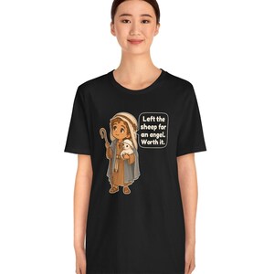 May include: A black t-shirt featuring a cartoon illustration of a shepherd holding a small white sheep. The shepherd wears a brown tunic and a blue cloak with a head covering. A speech bubble contains the text: Left the sheep for an angel. Worth it.