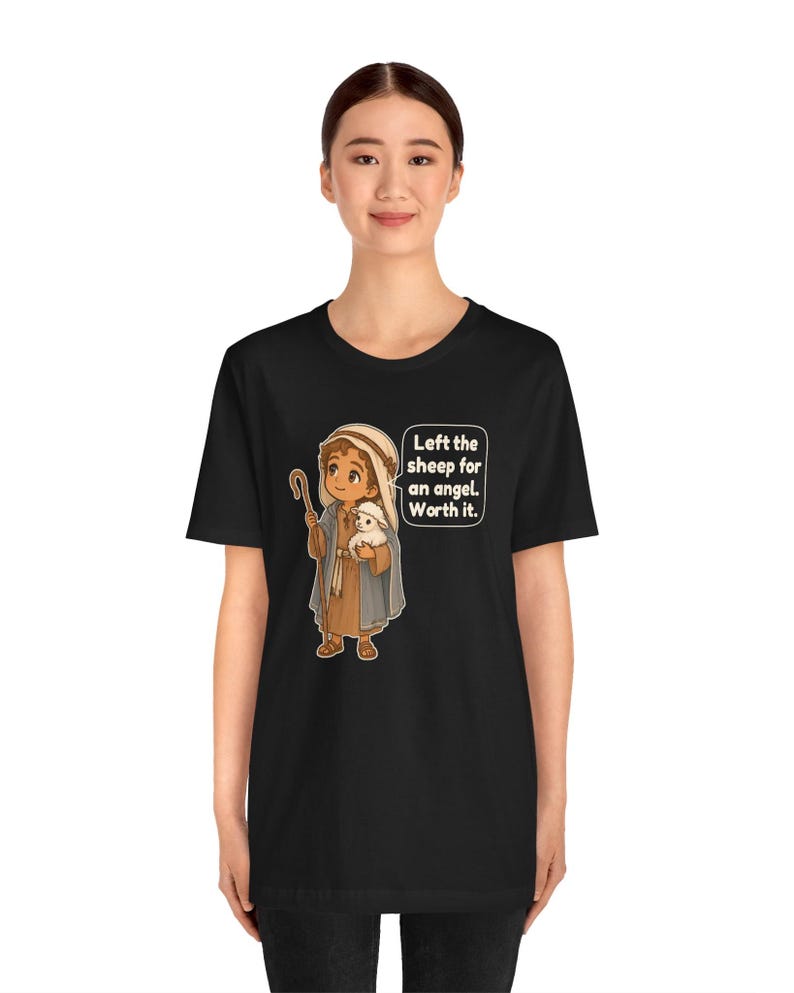 May include: A black t-shirt featuring a cartoon illustration of a shepherd holding a small white sheep. The shepherd wears a brown tunic and a blue cloak with a head covering. A speech bubble contains the text: Left the sheep for an angel. Worth it.