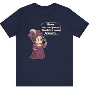 May include: A navy blue t-shirt featuring a cartoon illustration of a person in a purple Victorian-style dress and hat, holding a monocle. A speech bubble above the figure contains the text: Never learned piano. Would've been brilliant.