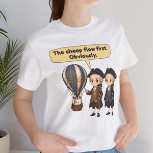 May include: A white t-shirt featuring a graphic of two figures in 18th-century attire standing next to a hot air balloon carrying a sheep. A speech bubble above them contains the text: The sheep flew first. Obviously.