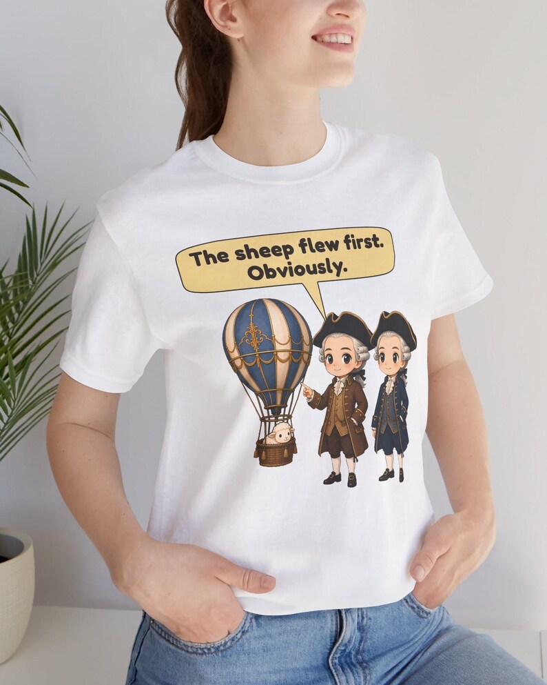 May include: A white t-shirt featuring a graphic of two figures in 18th-century attire standing next to a hot air balloon carrying a sheep. A speech bubble above them contains the text: The sheep flew first. Obviously.