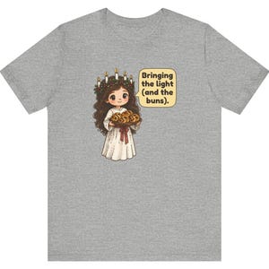 May include: A heather gray t-shirt featuring a cartoon illustration of a figure wearing a white gown and a candle crown, holding a tray of buns. A speech bubble next to the figure contains the text: Bringing the light (and the buns).