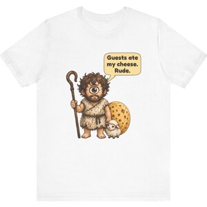 May include: A white t-shirt featuring a graphic of a cyclops wearing a fur tunic, holding a wooden staff, and standing next to a small sheep and a large wheel of cheese. A speech bubble above the cyclops reads, Guests ate my cheese. Rude.
