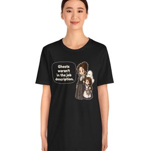 Op de afbeelding: Een zwart t-shirt met korte mouwen en een afbeelding van een vrouw en een kind met een pop, met een witte geest erachter. Een tekstballon bevat de tekst: Ghosts weren't in the job description.