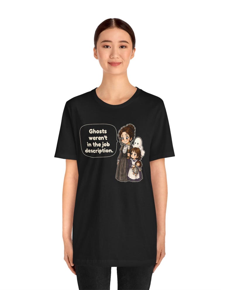 Op de afbeelding: Een zwart t-shirt met korte mouwen en een afbeelding van een vrouw en een kind met een pop, met een witte geest erachter. Een tekstballon bevat de tekst: Ghosts weren't in the job description.