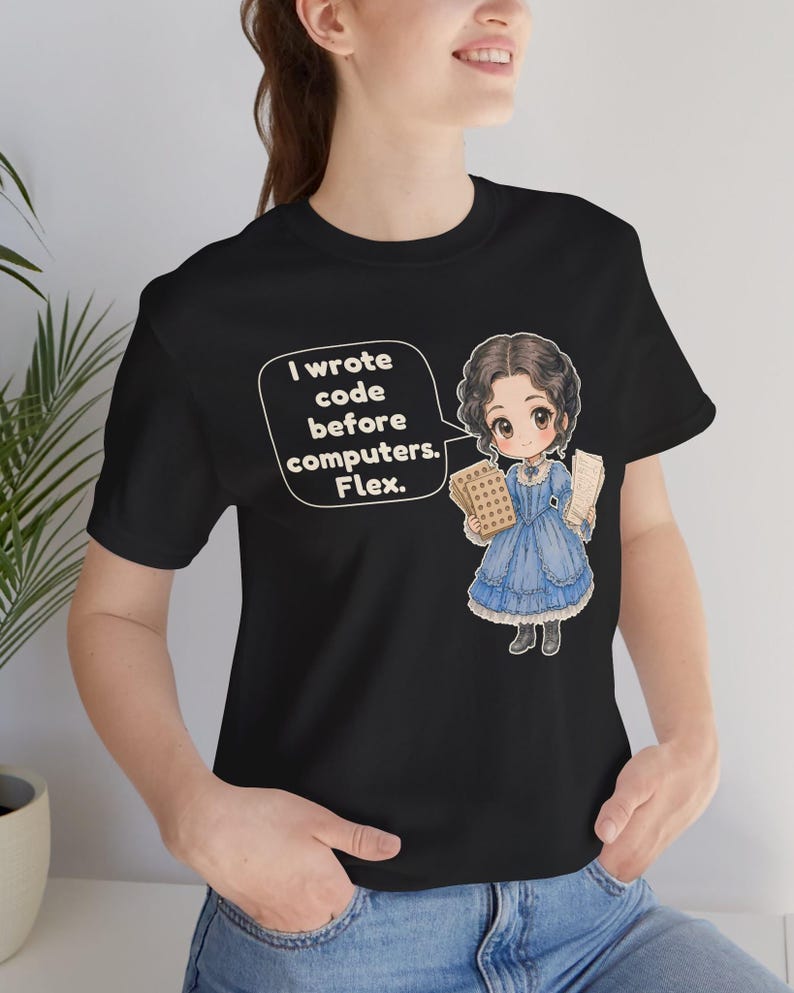 May include: A black t-shirt featuring a graphic of a person in a blue dress holding punch cards. A speech bubble next to the figure contains the text: I wrote code before computers. Flex. The design is centered on the front of the garment.