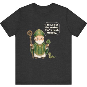 Puede incluir: Una camiseta gris jaspeado oscuro con una ilustraci&oacute;n de dibujos animados de una figura barbuda con vestiduras religiosas verdes, sosteniendo un bast&oacute;n de madera y un tr&eacute;bol, junto a una serpiente y un globo de texto: I drove out the snakes. You're next, Monday.