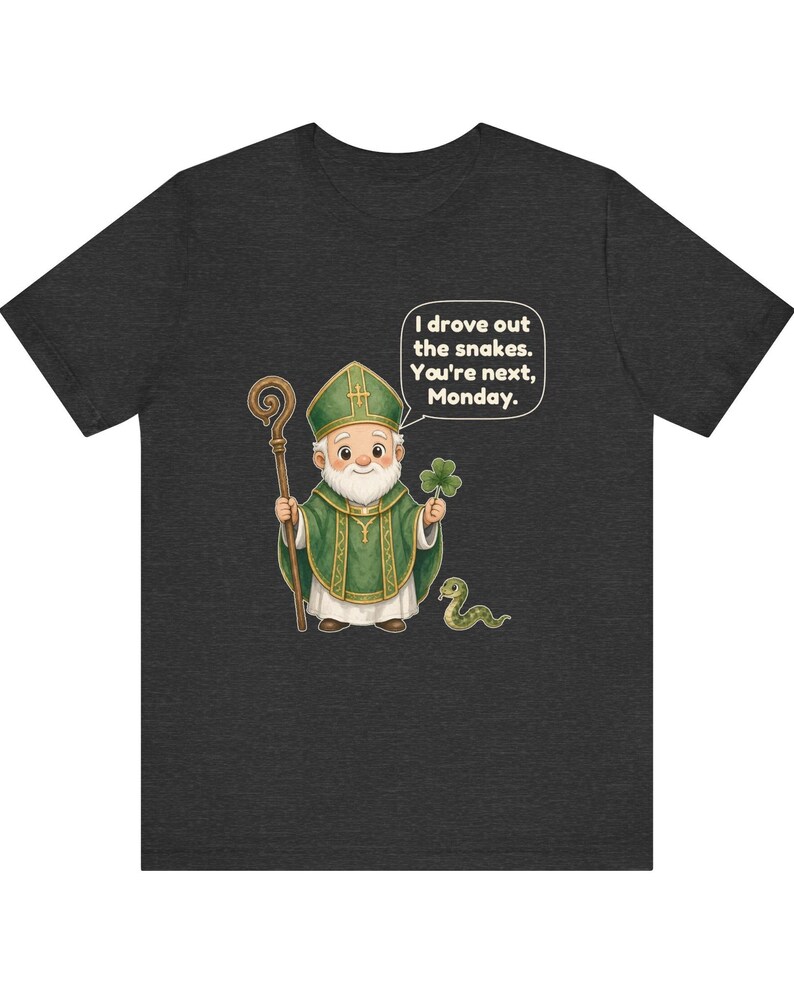 Puede incluir: Una camiseta gris jaspeado oscuro con una ilustraci&oacute;n de dibujos animados de una figura barbuda con vestiduras religiosas verdes, sosteniendo un bast&oacute;n de madera y un tr&eacute;bol, junto a una serpiente y un globo de texto: I drove out the snakes. You're next, Monday.