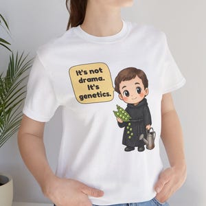 May include: A white short-sleeved t-shirt featuring a cartoon illustration of a monk holding green pea pods and a small watering can. A yellow speech bubble above the character contains the black text: It's not drama. It's genetics.