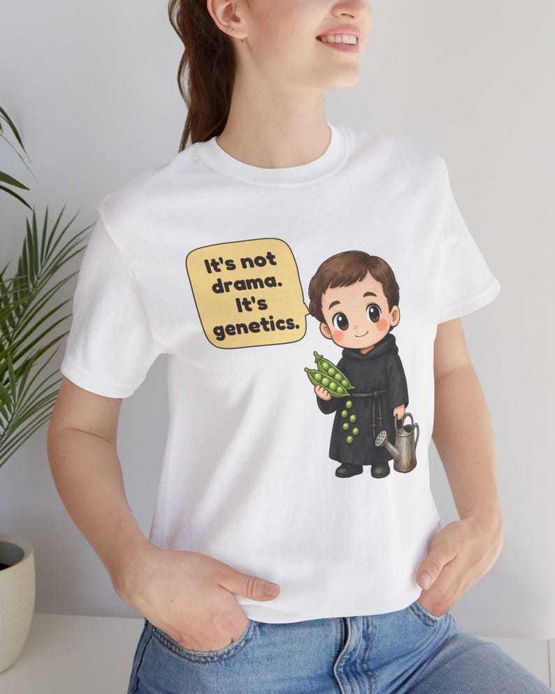 May include: A white short-sleeved t-shirt featuring a cartoon illustration of a monk holding green pea pods and a small watering can. A yellow speech bubble above the character contains the black text: It's not drama. It's genetics.