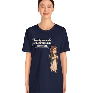 Op de afbeelding: Een marineblauw t-shirt met korte mouwen en een cartoonillustratie van een persoon in een lange jurk die een kaars vasthoudt. Een tekstballon boven het personage bevat de tekst: Twenty seconds of handwashing? Amateurs.
