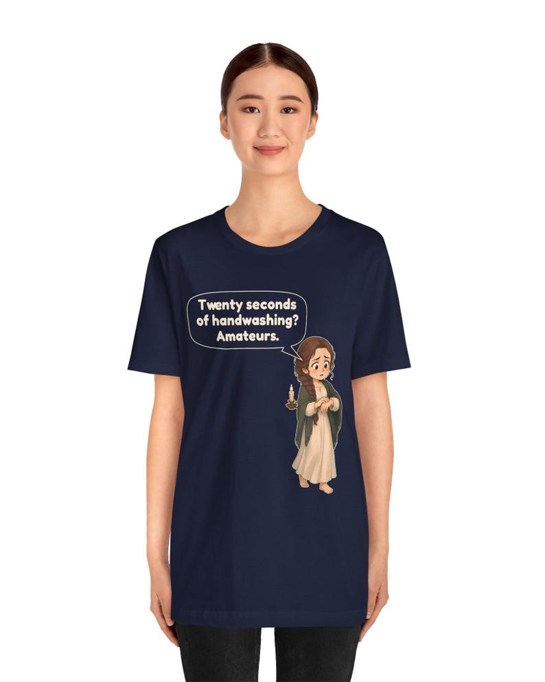 Op de afbeelding: Een marineblauw t-shirt met korte mouwen en een cartoonillustratie van een persoon in een lange jurk die een kaars vasthoudt. Een tekstballon boven het personage bevat de tekst: Twenty seconds of handwashing? Amateurs.