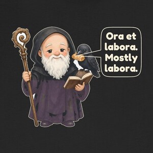 May include: A black t-shirt featuring a cartoon monk with a white beard and brown staff, holding an open book. A black raven sits on the book holding a piece of bread. A speech bubble contains the text: Ora et labora. Mostly labora.